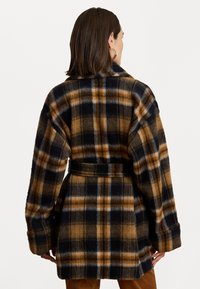 Cappotto di lana a quadri con una combinazione di colori marrone, nero e bianco, silhouette oversize, maniche ampie e una cintura coordinata in vita.