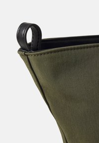 Grüne Stofftasche mit strukturierter Form, ausgestattet mit einem schwarzen Ledergriff und kontrastierendem Nähten entlang der Kanten. Texturierte Oberfläche.