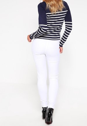 Femme portant un haut à manches longues rayé bleu marine et blanc, un pantalon blanc ajusté et des bottines noires à talons hauts, debout devant un fond uni.