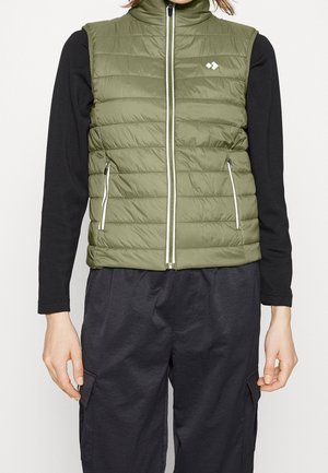 Person trägt eine ärmellose olivgrüne gesteppte Weste über einem schwarzen Langarmshirt und schwarze Cargohosen, steht mit entspannten Händen.