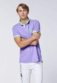 Lilafarbenes Poloshirt mit grünem Kragen und gestreiften Akzenten. Gesticktes Logo auf der Brust. Modell trägt weiße Hosen mit schwarzem Textdetail.