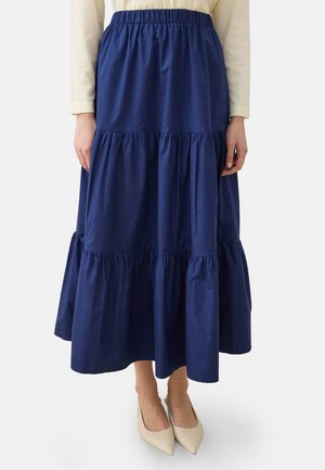 Gonna a balze di lunghezza media blu navy con vita elastica indossata con scarpe beige con punta appuntita e top crema a maniche lunghe, mostrata dalla vita in giù.