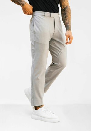 Άνδρας με ανοιχτό γκρι ραμμένο στα μέτρα παντελόνι, μαύρο πουκάμισο, λευκά αθλητικά παπούτσια, που δείχνει τατουαρισμένα χέρια και αστραγάλους σε λευκό φόντο.