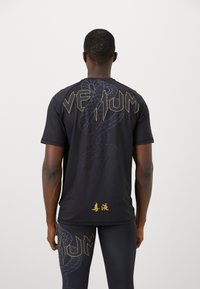 Hombre vestido con ropa deportiva negra con un gran gráfico de serpiente y el texto amarillo "Venum" en la parte posterior, junto con leggins a juego con diseño de serpiente.