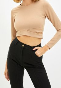 Langärmliges, verkürztes Oberteil aus beiger Stoffqualität mit tailliertem Schnitt. Kombiniert mit hochgeschnittenen schwarzen Hosen mit einem Knopf vorne und Taschen.