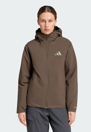 Kobieta ubrana w brązową kurtkę z kapturem i zamkiem błyskawicznym z logo Adidas, połączoną z ciemnymi spodniami, zwrócona przodem na jednolitym tle.