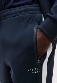 Χέρι στην τσέπη από σκούρο μπλε αθλητικό παντελόνι Ted Baker με λευκή πλαϊνή ρίγα και λεπτομέρεια με φερμουάρ στην τσέπη.