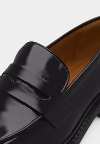 Μαύρο γυαλιστερό δερμάτινο μοκασίνι με λουράκι πάνω από το επάνω μέρος και εσωτερική επένδυση σε ταμπά χρώμα.