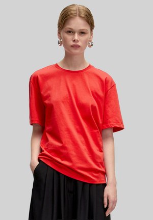 Rode katoenen T-shirt met ronde hals en korte mouwen, gepresenteerd op een model, gecombineerd met een zwarte rok. Gladde textuur en casual pasvorm.