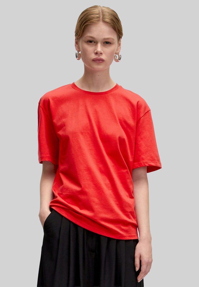 Rotes Baumwoll-T-Shirt mit Rundhalsausschnitt und kurzen Ärmeln, auf einem Model präsentiert, kombiniert mit einem schwarzen Rock. Weiche Textur und lässige Passform.