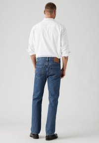 Jeans in denim blu classico con vestibilità dritta, dotati di un'etichetta in pelle sulla parte posteriore e tasche cucite. Indossati con una camicia bianca.