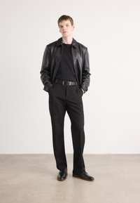 Veste en cuir noir, pull noir et pantalon noir ajusté, complétés par des chaussures noires avec des semelles brunes, sur un fond clair.