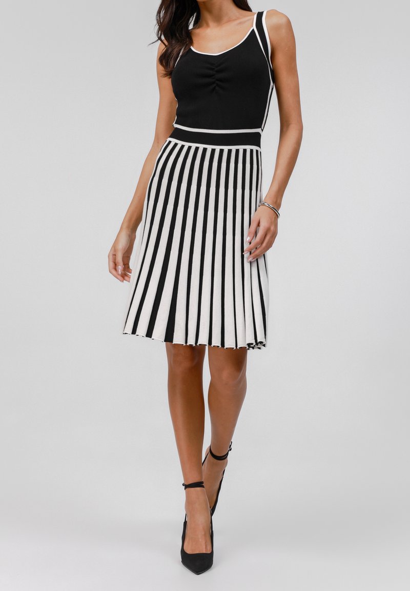 Femme portant un haut noir sans manches avec un bord blanc et une jupe plissée noire et blanche à rayures, longueur genou, associée à des talons noirs.