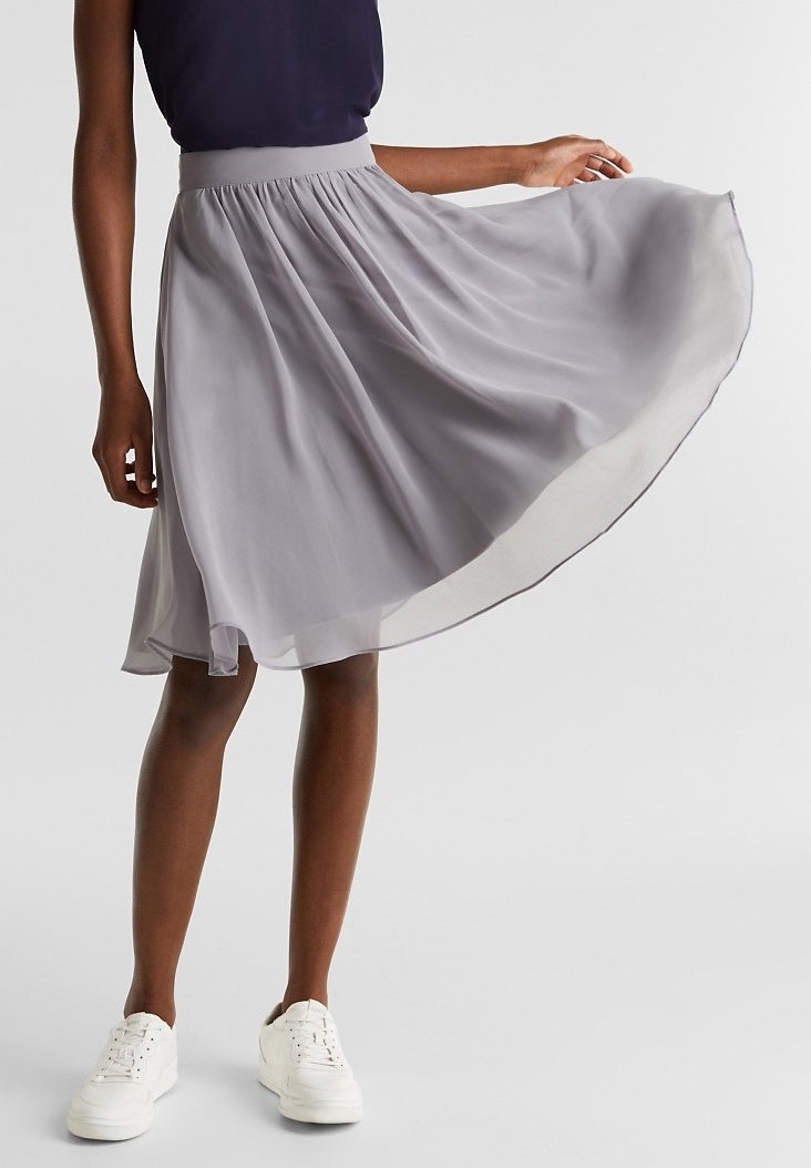 Femme portant une jupe fluide gris clair à la hauteur des genoux, un top sans manches bleu marine et des baskets blanches, tenant la jupe sur le côté.