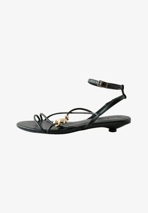 Black flat sandal with thin straps, gold decorative bead on front strap, and ankle buckle closure.