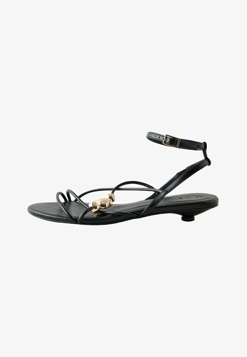 Sandale plate noire avec fines lanières, perle décorative dorée sur la lanière avant, et fermeture à boucle à la cheville.