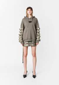 Sudadera gris de gran tamaño con detalles metálicos en las mangas, combinada con pantalones cortos metálicos. La modelo lleva tacones negros de punta afilada y una gorra a juego.