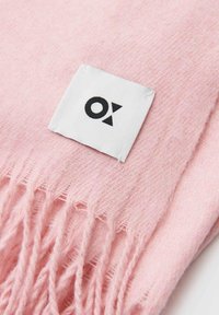 Écharpe en laine rose avec bords à franges, dotée d'une étiquette blanche avec un logo noir. Texture douce et design léger.