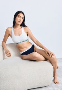 Witte sportbeha met een groen en marineblauwe gestreepte band en een bijpassende marineblauwe bikini met een witte afwerking, zittend op een gestructureerde beige bank.