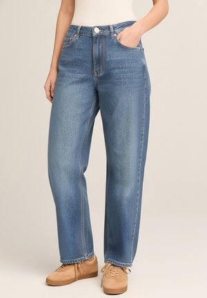 Woman wearing straight-leg blue jeans, beige sneakers, and a white top, standing with one hand in pocket against plain background.