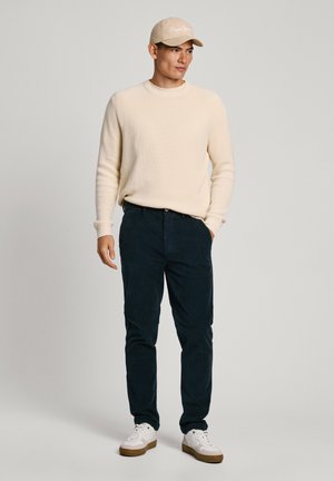 Maglione beige a coste, pantaloni in corduroy blu, sneakers bianche con suole marroni e un cappellino beige con testo a contrasto. Posizione eretta su uno sfondo neutro.