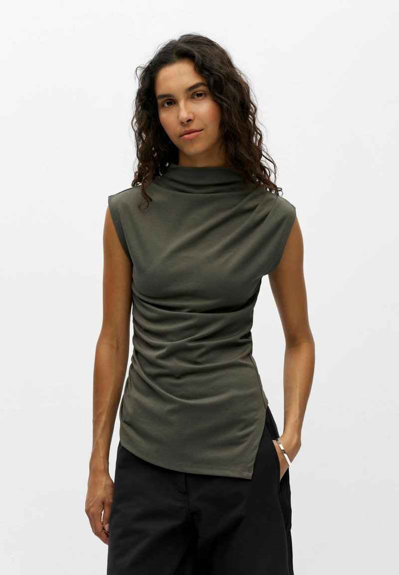 Femme aux cheveux bouclés et foncés portant un haut sans manches vert olive et un pantalon noir, debout avec une main dans la poche devant un fond uni.