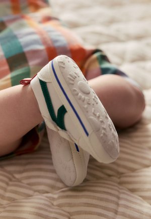 Bébé portant des chaussures blanches à semelle souple avec des accents verts et bleus, couché sur une surface rayée avec des vêtements à carreaux colorés visibles.