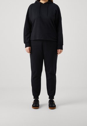 Personne portant un sweat à capuche noir, un pantalon de jogging noir et des baskets noires avec des semelles marron, debout devant un fond clair uni.