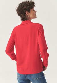 Blusa roja de manga larga con cuello, textura suave y detalles de botones en los puños. Se muestra desde la parte posterior, combinada con jeans de mezclilla azules.
