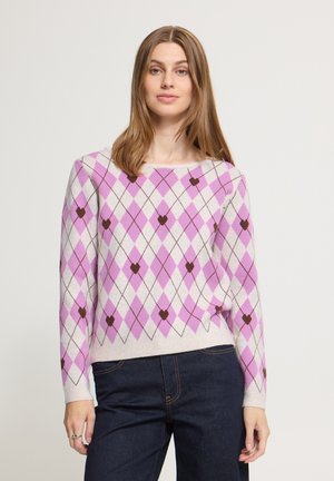 Vrouw staat frontaal, draagt een lichtpaarse en witte argyle trui met bruine hartvormen, gecombineerd met donkerblauwe jeans. Neutrale achtergrond.