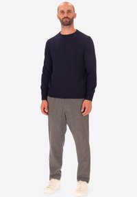Uomo con barba che indossa un maglione blu navy, pantaloni grigi e sneakers bianche, in piedi di fronte a uno sfondo chiaro e neutro.