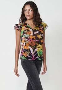 Blusa nera trasparente con stampa floreale colorata, maniche a sbuffo arricciate e scollatura a V. Abbinata a jeans scuri.