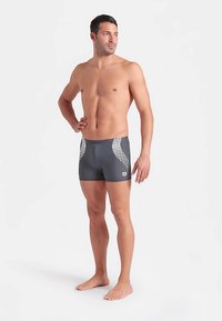 Grå badebukser med en tætsiddende pasform, der har et tekstureret hvidt mønster på siderne. Glat stof og lavtaljet linning.