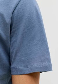 Nära bild av en blå kortärmad bomulls t-shirt på en persons arm mot en enkel ljus bakgrund.