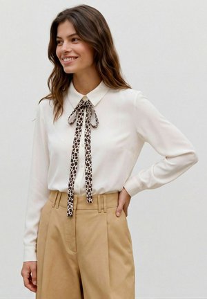 Femme souriante portant un chemisier blanc à manches longues avec un nœud papillon à imprimé léopard et un pantalon beige taille haute, main sur la hanche.