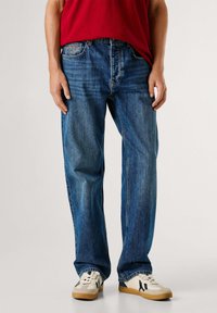 Vaqueros de mezclilla azul con un corte recto, que presentan un diseño clásico de cinco bolsillos y una etiqueta con el logo de cuero color marrón claro en la parte trasera. Combinados con zapatillas blancas.