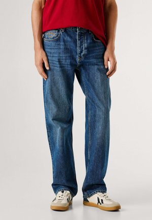 Jean en denim bleu à coupe droite, doté d'un design classique à cinq poches et d'un patch logo en cuir beige à l'arrière. Porté avec des baskets blanches.