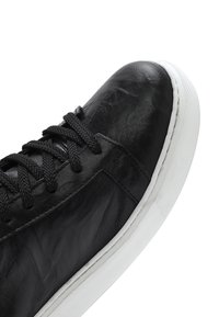 Zwarte leren sneaker met zwarte veters en dikke witte rubberen zool, gezien vanuit een boven-zijaanzicht op een witte achtergrond.