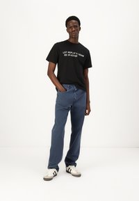 Svart T-skjorte med hvit tekst, kombinert med blå løstsittende jeans. Modellen har på seg hvite og grå joggesko med svarte striper. Enkelt design, uformell stil.