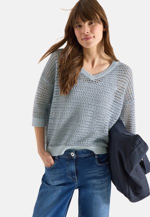 Vrouw met lang bruin haar, gekleed in een lichtgrijze, opengebreide trui en een blauwe spijkerbroek, die een donker stoffen tas in haar linkerhand vasthoudt.