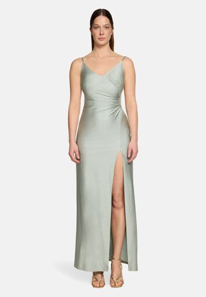 Kobieta w jasnooliwkowej, bezrękawnikowej sukni sięgającej podłogi z wysokim rozcięciem na udzie, marszczonym pasem i złotymi sandałami na cienkich paskach, stojąca na jednolitym tle.