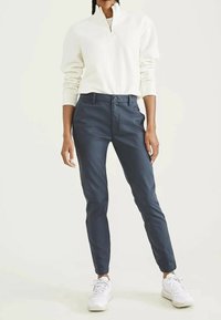 Person som bär en vit zip-up tröja, marinblå slim-fit byxor och vita sneakers som står mot en enkel ljus bakgrund.