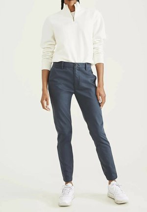 Persoon in witte quarter-zip trui, marineblauwe slim-fit broek en witte sneakers, staand tegen een egale lichte achtergrond.