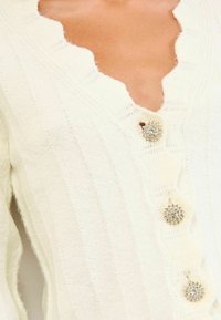 Közelkép egy fehér kötött kardigánról, hullámos nyakkivágással és díszes gyöngy gombokkal az elején.