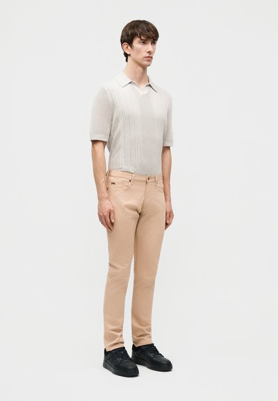 Νεαρός άνδρας όρθιος φορώντας ένα μπεζ πλεκτό πόλο με κοντά μανίκια, ανοιχτό καφέ στενά παντελόνια και μαύρα αθλητικά παπούτσια σε απλό ανοιχτό φόντο.