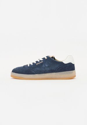 Navy blue suede sneakers with a gum rubber sole, white heel accent, and beige laces. Embossed logo on the side. Low-profile design.