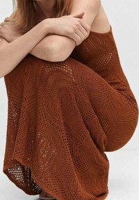 Femme assise avec les bras enroulés autour des genoux, portant une robe en croche rustique avec des motifs complexes.