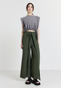 Frau in grauem ärmellosem Crop-Top mit geraffter Taille, weit geschnittenen olivgrünen Hosen mit Bindegürtel und schwarzen Plateau-Sandalen, die auf einem weißen Boden steht.