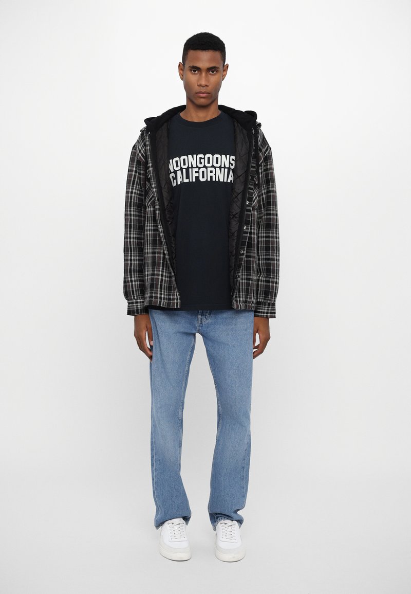 Veste à carreaux noire et grise portée sur un T-shirt noir avec un texte blanc, jean bleu clair et baskets blanches. Le mannequin se tient devant un fond uni.