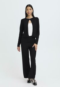 Schwarze, kürzere Strickjacke mit einem Ausschnitt vorne, kombiniert mit schwarzen schmalen Hosen und schwarzen Schuhen mit einer strukturierten Akzentuierung.